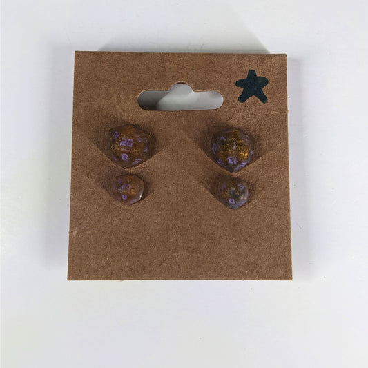 Two pairs of handmade d20 earrings with sterling silver hardware that are dark gold with purple numbers on a kraft brown earring card with a star in the top right corner. The earrings on the left of each pair centers the 20 face and the earrings on the right of each pair centers the 1 face.