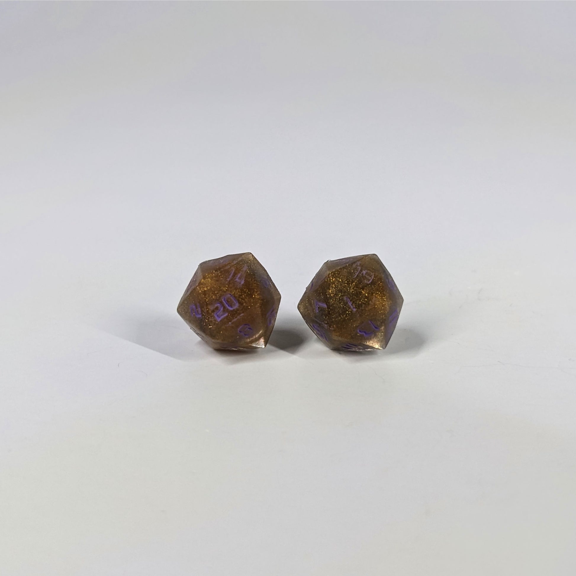 A single pair of the larger size handmade d20 earrings with sterling silver hardware that are dark gold with purple numbers on a white background. The earring on the left centers the 20 face and the earring on the right centers the 1 face.