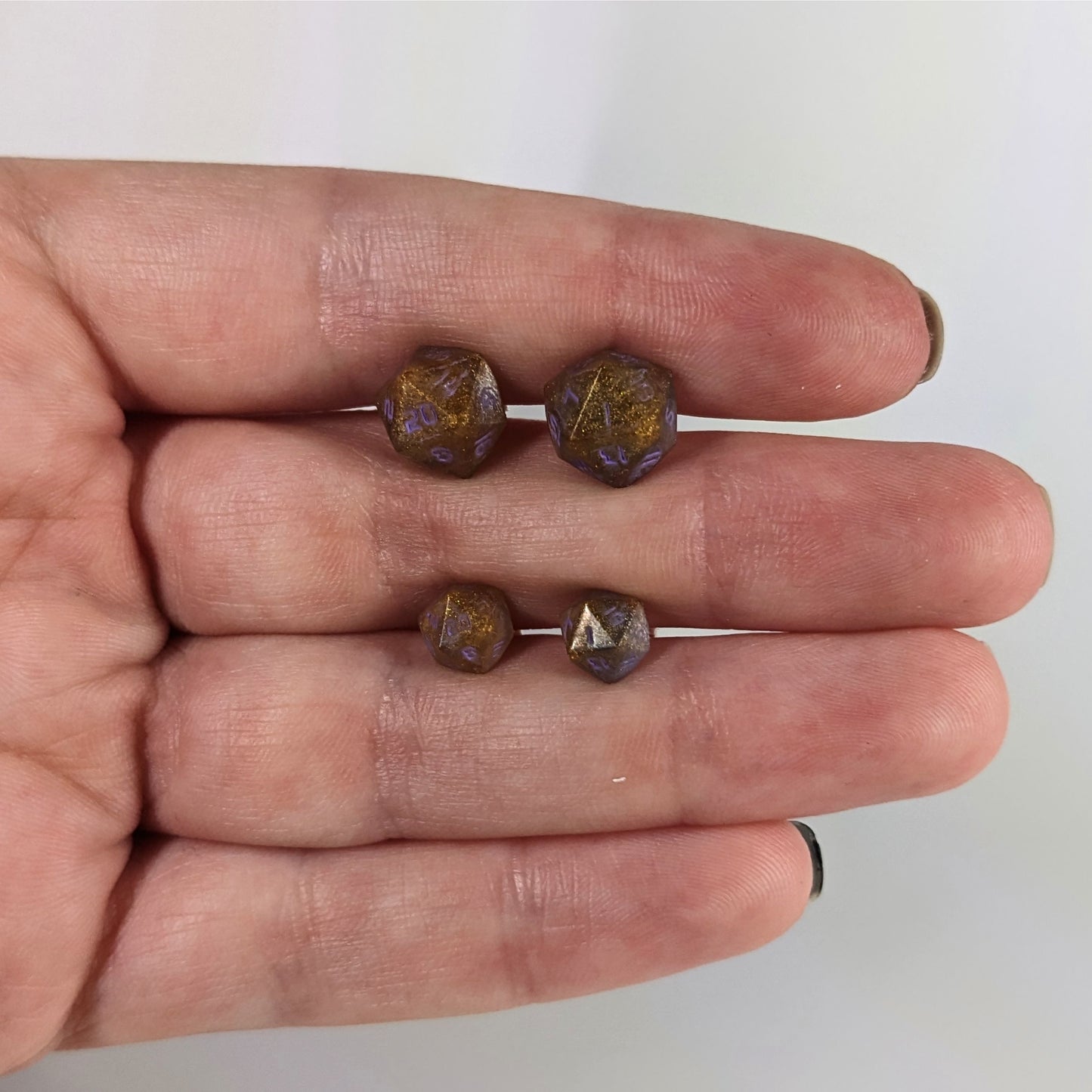 Two pairs of handmade d20 earrings with sterling silver hardware that are dark gold with purple numbers held between the fingers of a flat hand. The larger pair is between the pointer and middle finger, the smaller between the middle and ring finger. The earrings on the left of each pair centers the 20 face and the earrings on the right of each pair centers the 1 face.