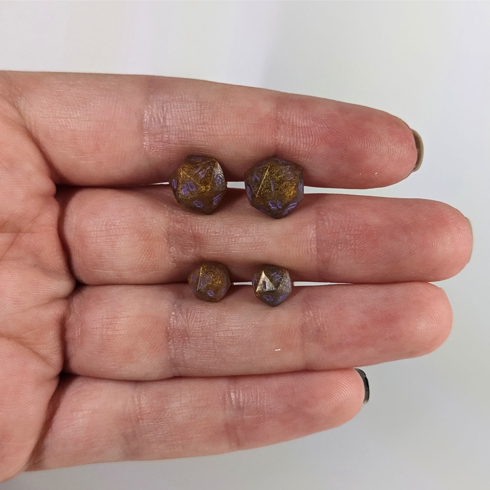 Two pairs of handmade d20 earrings with sterling silver hardware that are dark gold with purple numbers held between the fingers of a flat hand. The larger pair is between the pointer and middle finger, the smaller between the middle and ring finger. The earrings on the left of each pair centers the 20 face and the earrings on the right of each pair centers the 1 face.