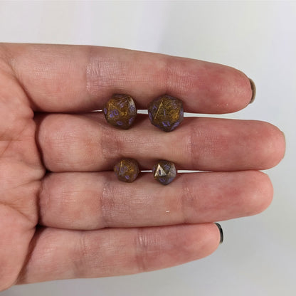 Two pairs of handmade d20 earrings with sterling silver hardware that are dark gold with purple numbers held between the fingers of a flat hand. The larger pair is between the pointer and middle finger, the smaller between the middle and ring finger. The earrings on the left of each pair centers the 20 face and the earrings on the right of each pair centers the 1 face.