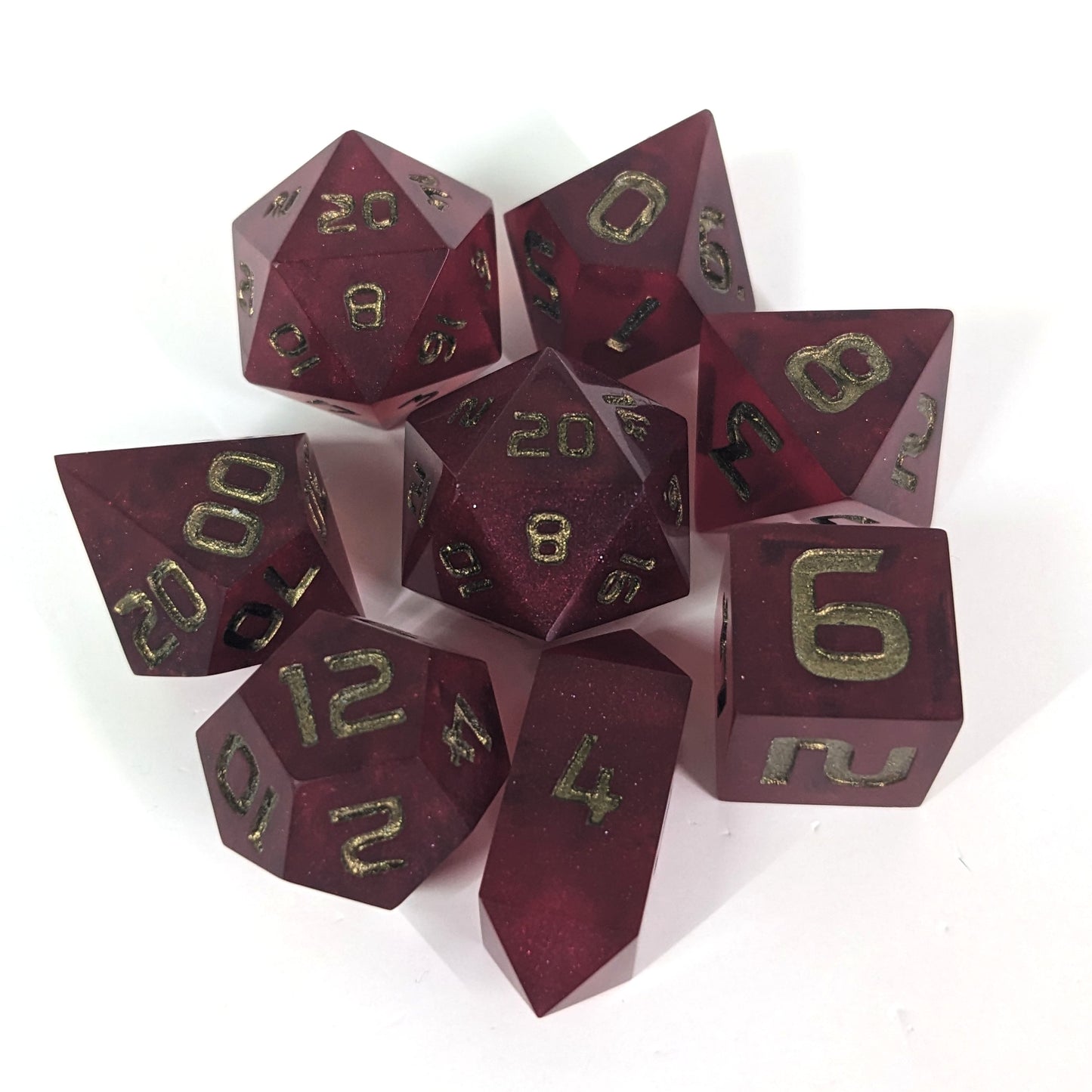 A complete set of 8 handmade artisan dice that are bold red with bronze numbers. The DnD dice set is arranged in a circle and features 2 D20s, one of which is in the center surrounded by the other 7 dice on a white background. The d4 is a unique, crystalline shape.