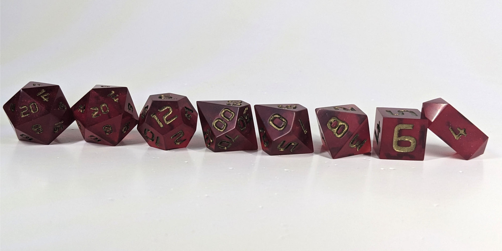 A straight on photo of a complete set of polyhedral dice that are bold red with bronze numbers arranged in a straight line. They are lined up with the 2 d20s on the left going down to the d4 on the right side. The highest value number of each die is facing forward, and the crystalline d4 is leaning against the d6.