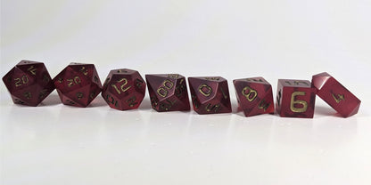 A straight on photo of a complete set of polyhedral dice that are bold red with bronze numbers arranged in a straight line. They are lined up with the 2 d20s on the left going down to the d4 on the right side. The highest value number of each die is facing forward, and the crystalline d4 is leaning against the d6.