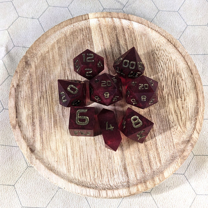 A complete set of 8 handmade artisan dice that are a bold red with bronze numbers. The DnD dice set is on a wooded circle and the dice are arranged in a circle and features 2 D20s, one of which is in the center surrounded by the other 7 dice. The d4 is unique with a crystalline shape.