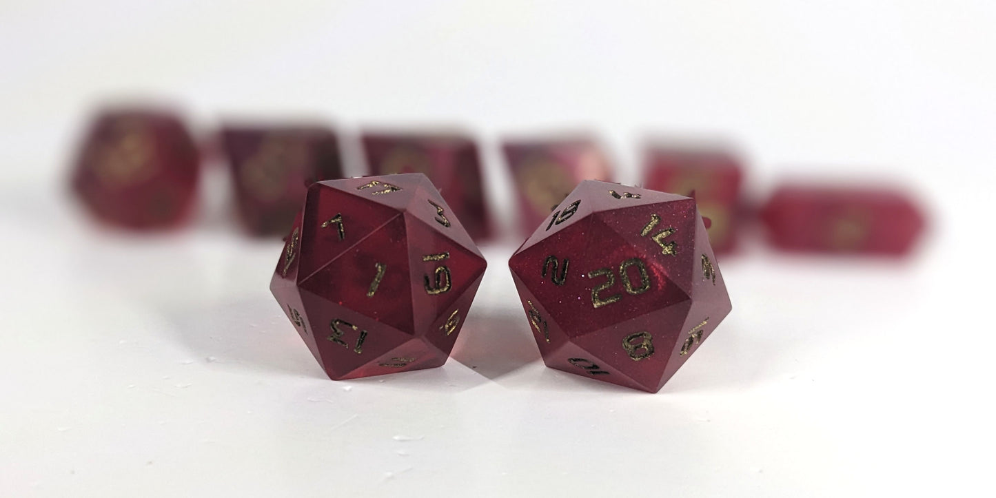 A straight on photo of a standard set of DnD dice that are bold red with bronze numbers focusing on 2 d20s in the center. The d20 on the left has the 1 face centered and the d20 on the right has the 20 face centered. The other 6 dice are lined up in the background but are out of focus, while the d20s are in sharp focus.
