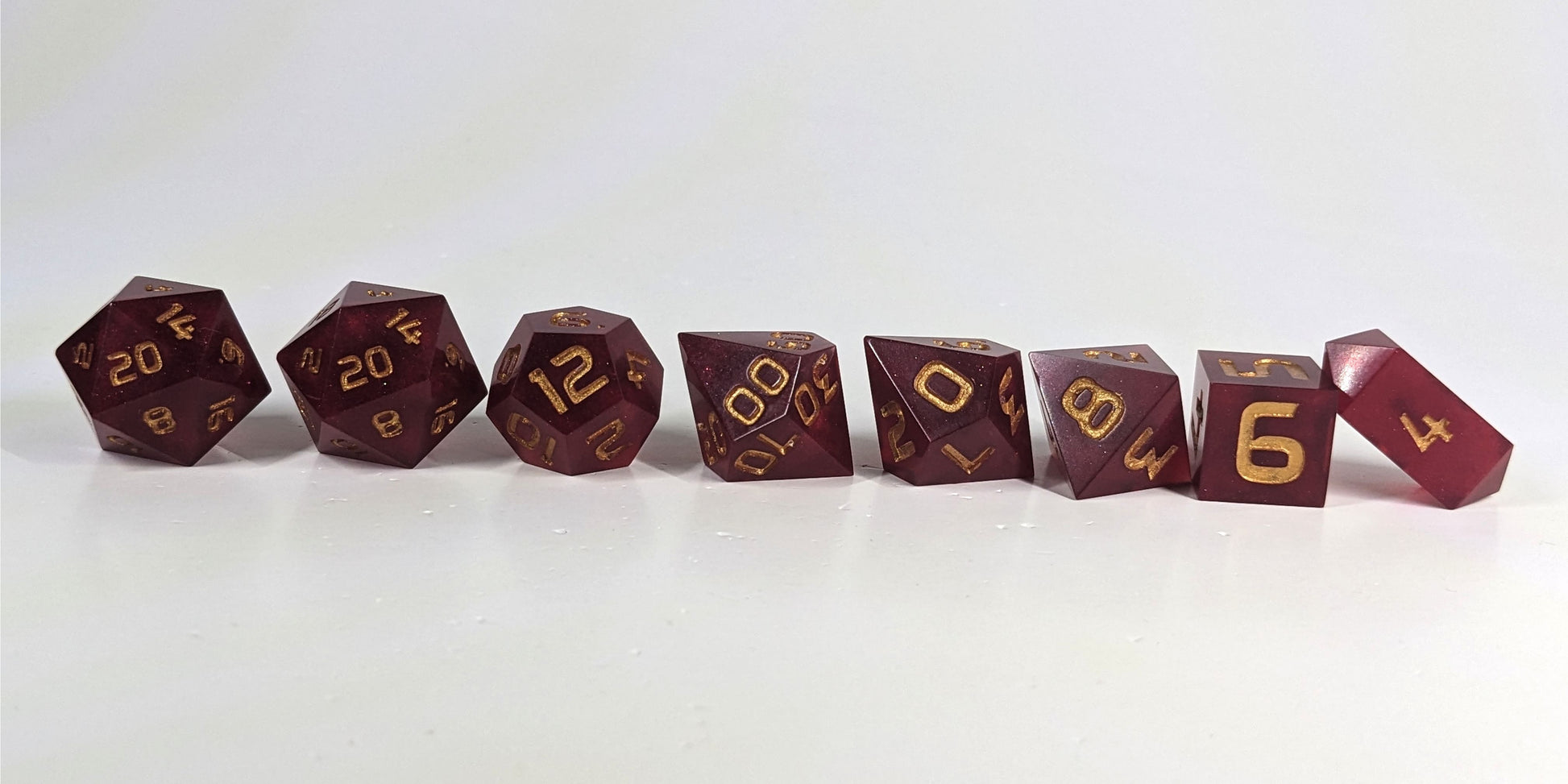 A straight on photo of a complete set of polyhedral dice that are bold red with gold numbers arranged in a straight line. They are lined up with the 2 d20s on the left going down to the d4 on the right side. The highest value number of each die is facing forward, and the crystalline d4 is leaning against the d6.