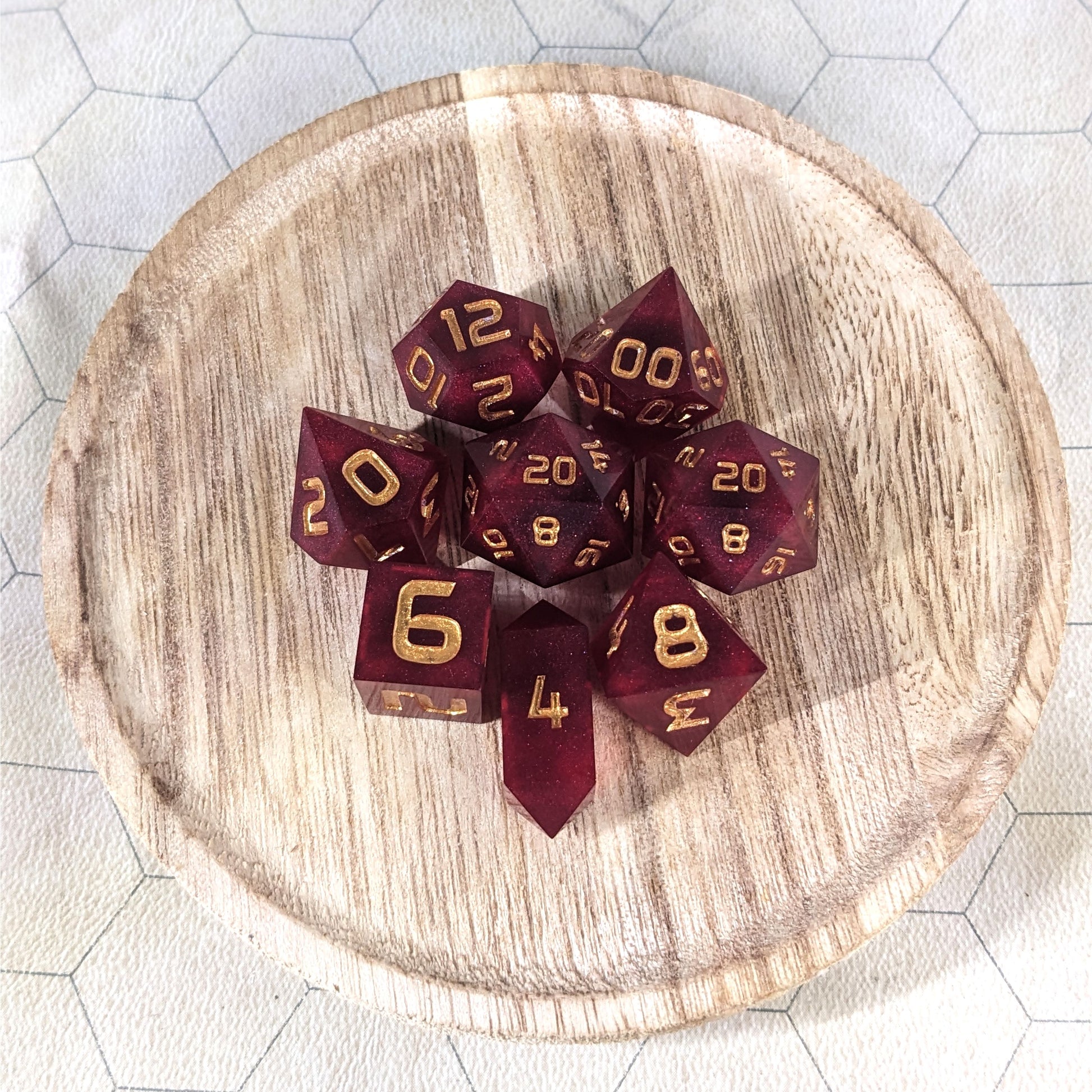 A complete set of 8 handmade artisan dice that are a bold red with gold numbers. The DnD dice set is on a wooded circle and the dice are arranged in a circle and features 2 D20s, one of which is in the center surrounded by the other 7 dice. The d4 is unique with a crystalline shape.
