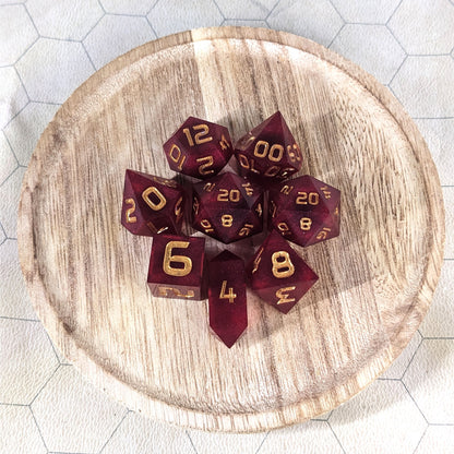 A complete set of 8 handmade artisan dice that are a bold red with gold numbers. The DnD dice set is on a wooded circle and the dice are arranged in a circle and features 2 D20s, one of which is in the center surrounded by the other 7 dice. The d4 is unique with a crystalline shape.
