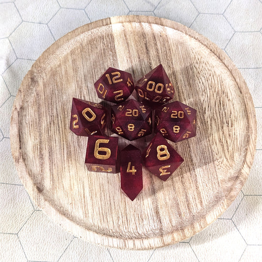 A complete set of 8 handmade artisan dice that are a bold red with gold numbers. The DnD dice set is on a wooded circle and the dice are arranged in a circle and features 2 D20s, one of which is in the center surrounded by the other 7 dice. The d4 is unique with a crystalline shape.