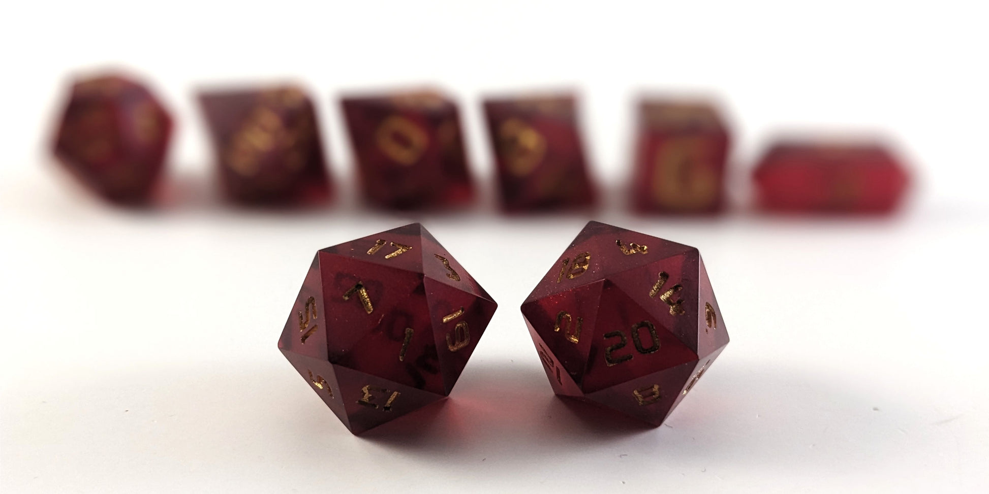 A straight on photo of a standard set of DnD dice that are bold red with gold numbers focusing on 2 d20s in the center. The d20 on the left has the 1 face centered and the d20 on the right has the 20 face centered. The other 6 dice are lined up in the background but are out of focus, while the d20s are in sharp focus.