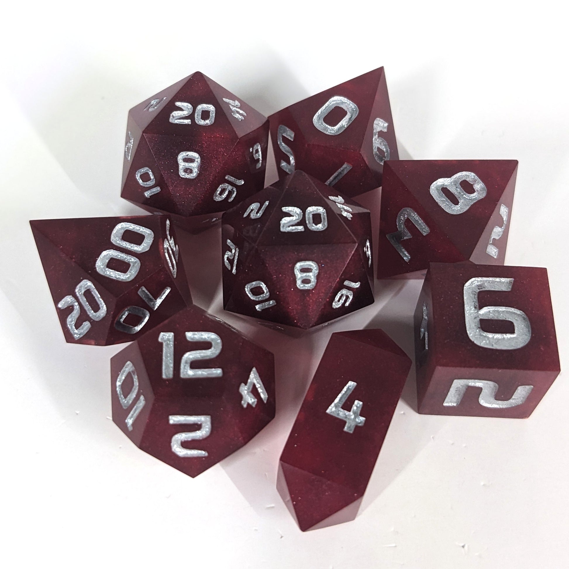 A complete set of 8 handmade artisan dice that are bold red with silver numbers. The DnD dice set is arranged in a circle and features 2 D20s, one of which is in the center surrounded by the other 7 dice on a white background. The d4 is a unique, crystalline shape.