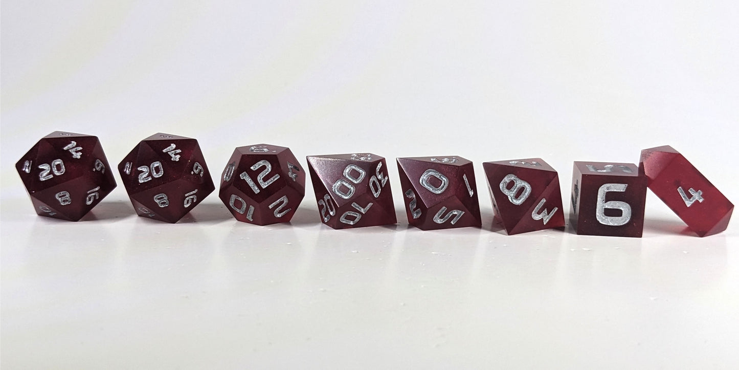 A straight on photo of a complete set of polyhedral dice that are bold red with silver numbers arranged in a straight line. They are lined up with the 2 d20s on the left going down to the d4 on the right side. The highest value number of each die is facing forward, and the crystalline d4 is leaning against the d6.