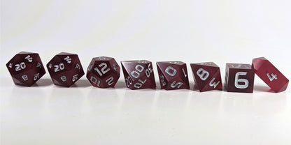 A straight on photo of a complete set of polyhedral dice that are bold red with silver numbers arranged in a straight line. They are lined up with the 2 d20s on the left going down to the d4 on the right side. The highest value number of each die is facing forward, and the crystalline d4 is leaning against the d6.