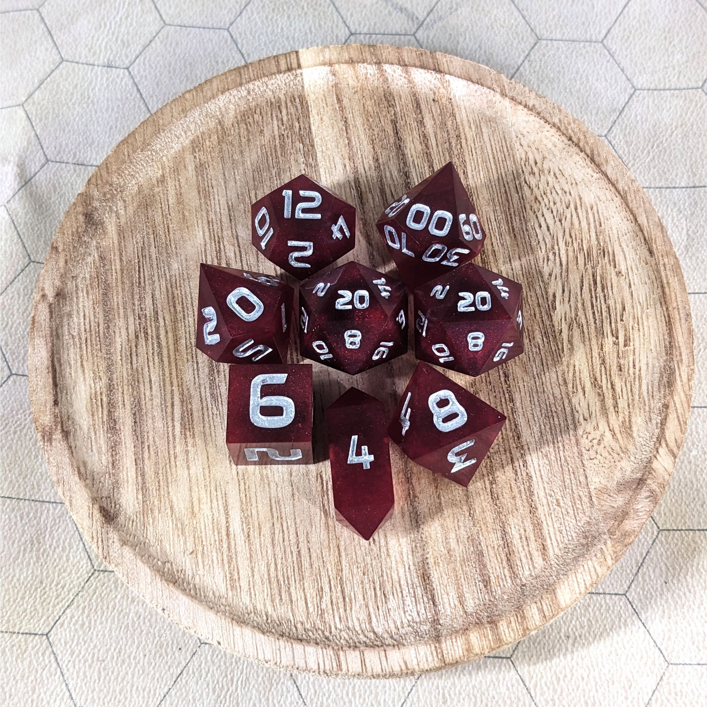 A complete set of 8 handmade artisan dice that are a bold red with silver numbers. The DnD dice set is on a wooded circle and the dice are arranged in a circle and features 2 D20s, one of which is in the center surrounded by the other 7 dice. The d4 is unique with a crystalline shape.