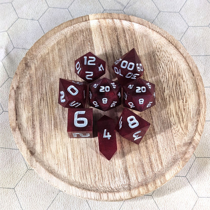 A complete set of 8 handmade artisan dice that are a bold red with silver numbers. The DnD dice set is on a wooded circle and the dice are arranged in a circle and features 2 D20s, one of which is in the center surrounded by the other 7 dice. The d4 is unique with a crystalline shape.
