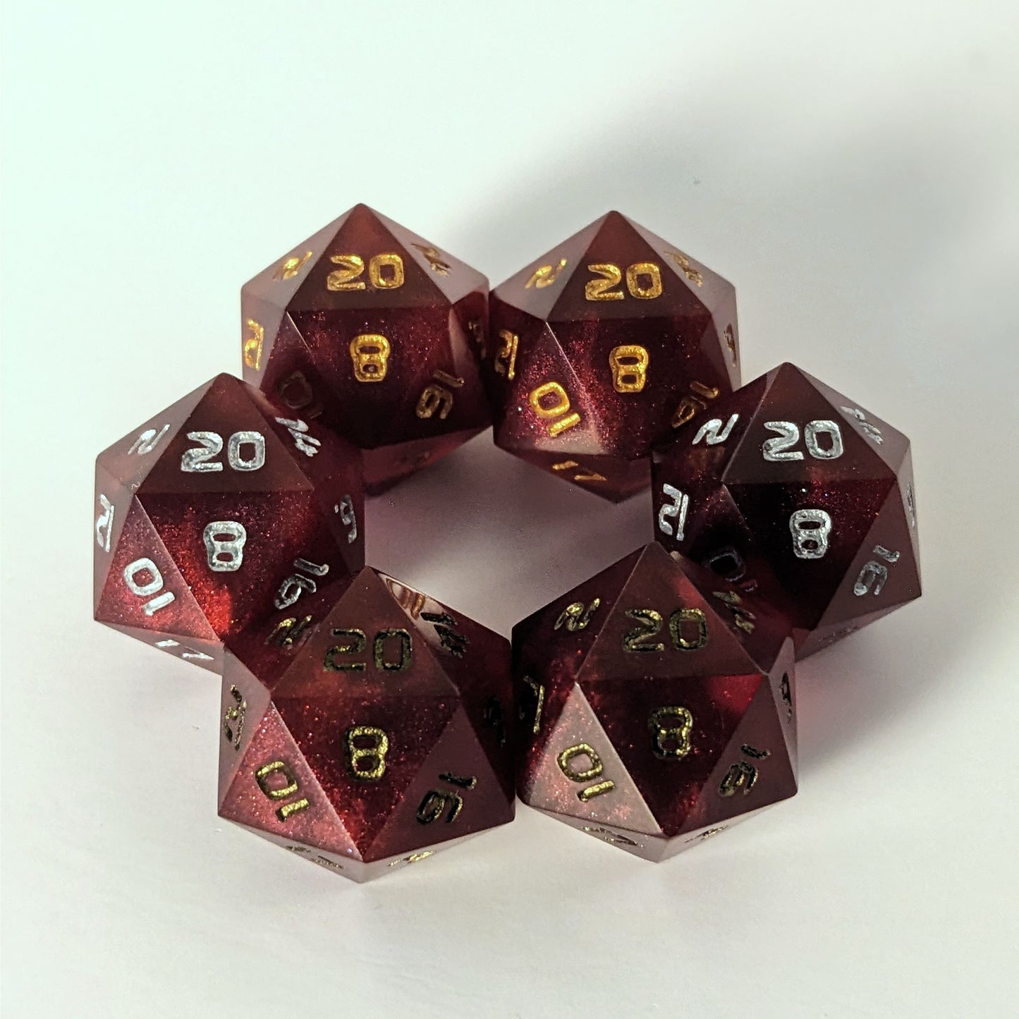 Three pairs of standard d20s are arranged in a hexagon on a white background. All of the handmade dice are a bold red. The top two d20s have gold numbers, the middle two have silver numbers, and the bottom two have bronze numbers, showing how each number color affects the appearance of the hue of the crimson red dice.