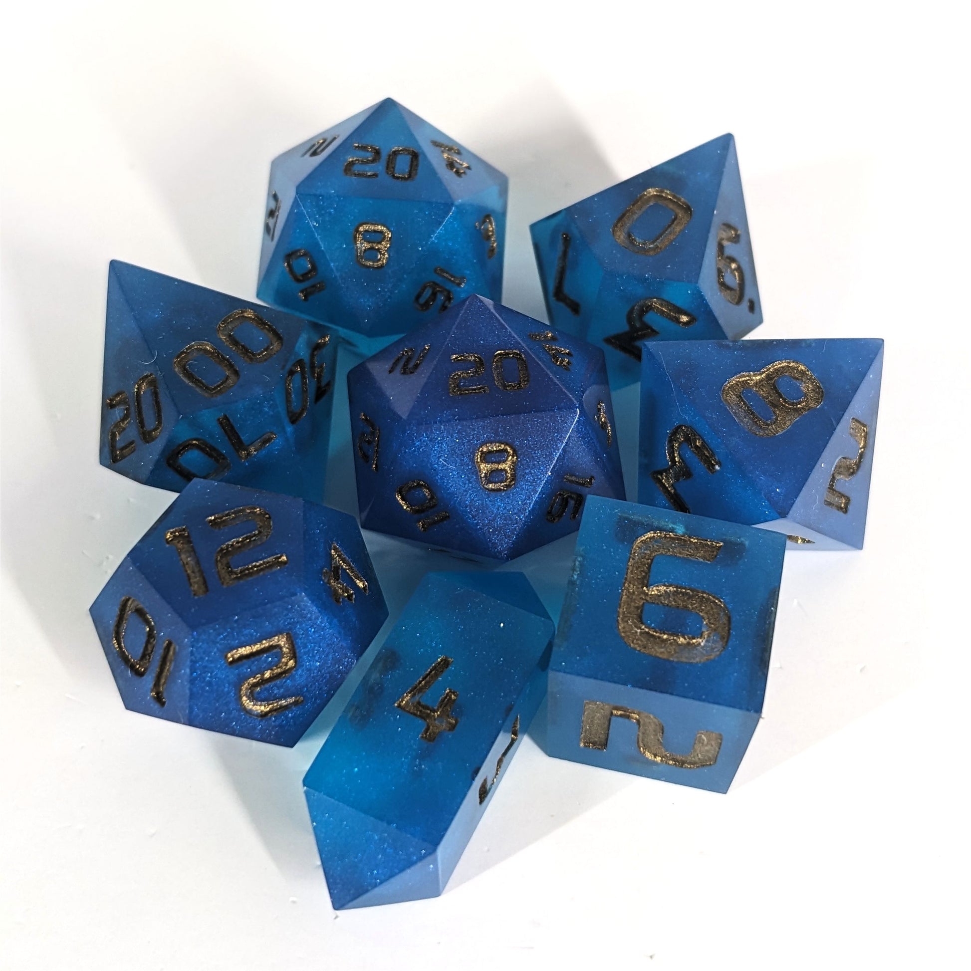 A complete set of 8 handmade artisan dice that are rich blue with bronze numbers. The DnD dice set is arranged in a circle and features 2 D20s, one of which is in the center surrounded by the other 7 dice on a white background. The d4 is a unique, crystalline shape.