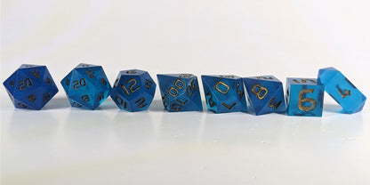 A straight on photo of a complete set of polyhedral dice that are rich blue with bronze numbers arranged in a straight line. They are lined up with the 2 d20s on the left going down to the d4 on the right side. The highest value number of each die is facing forward, and the crystalline d4 is leaning against the d6.