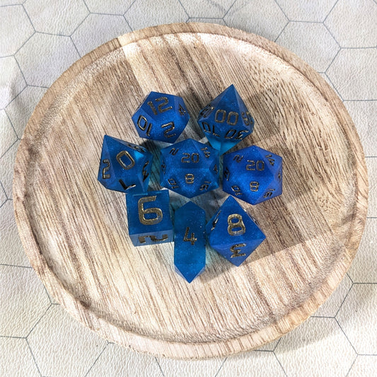 A complete set of 8 handmade artisan dice that are a rich blue with bronze numbers. The DnD dice set is on a wooded circle and the dice are arranged in a circle and features 2 D20s, one of which is in the center surrounded by the other 7 dice. The d4 is unique with a crystalline shape.