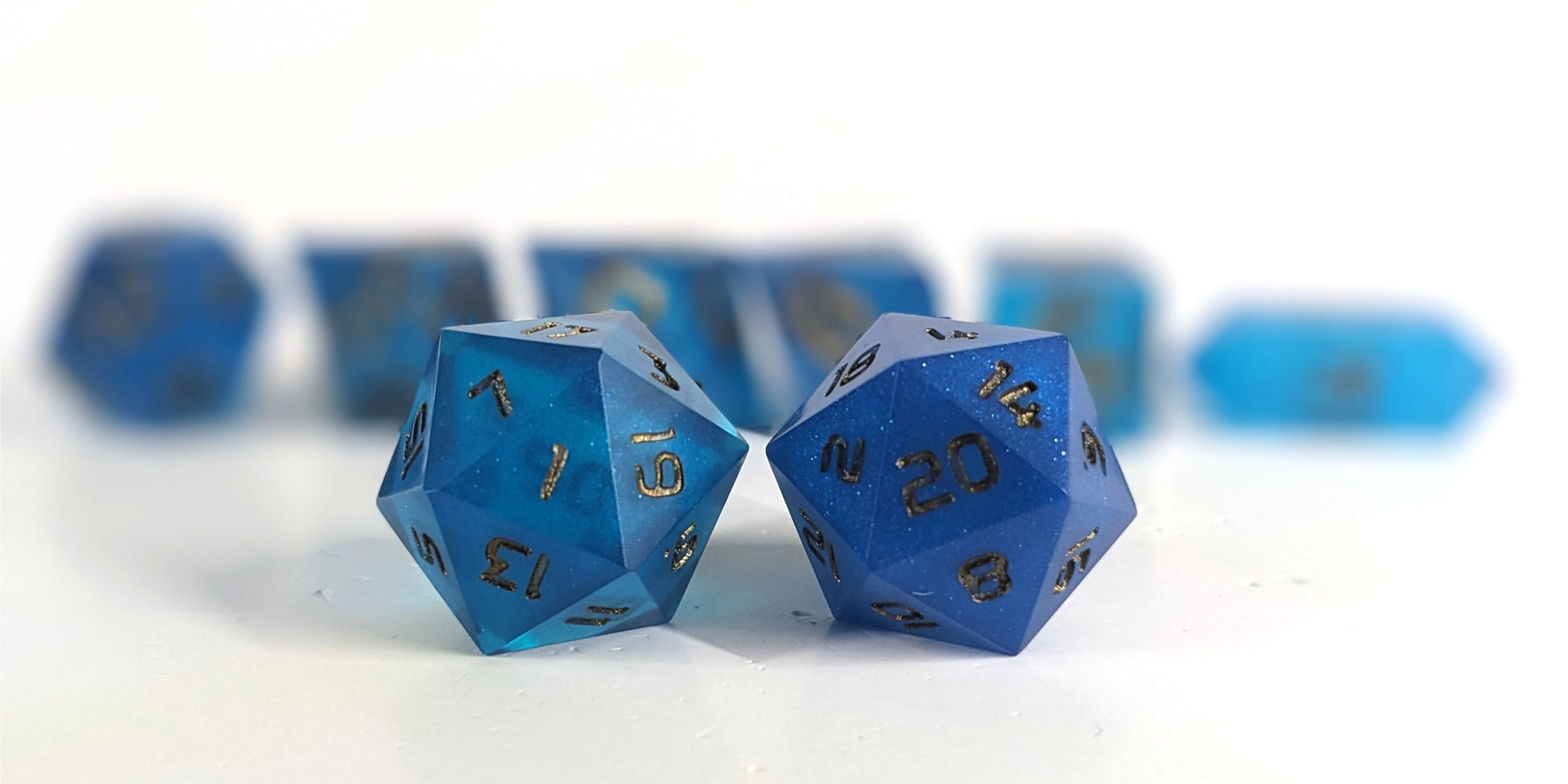 A straight on photo of a standard set of DnD dice that are rich blue with bronze numbers focusing on 2 d20s in the center. The d20 on the left has the 1 face centered and the d20 on the right has the 20 face centered. The other 6 dice are lined up in the background but are out of focus, while the d20s are in sharp focus.