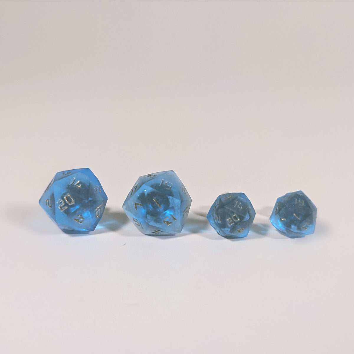 Two pairs of handmade d20 earrings with titanium hardware that are sapphire blue with bronze numbers on a white background sit in a line with the larger pair on the left and the smaller pair on the right. The earrings on the left of each pair centers the 20 face and the earrings on the right of each pair centers the 1 face.