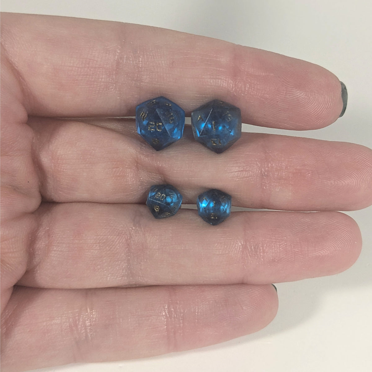 Two pairs of handmade d20 earrings with titanium hardware that are sapphire blue with bronze numbers held between the fingers of a flat hand. The larger pair is between the pointer and middle finger, the smaller between the middle and ring finger. The earrings on the left of each pair centers the 20 face and the earrings on the right of each pair centers the 1 face.