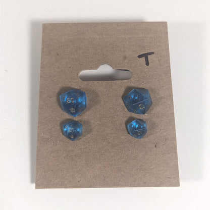 Two pairs of handmade d20 earrings with titanium hardware that are sapphire blue with bronze numbers on a kraft brown earring card with a T in the top right corner. The earrings on the left of each pair centers the 20 face and the earrings on the right of each pair centers the 1 face.