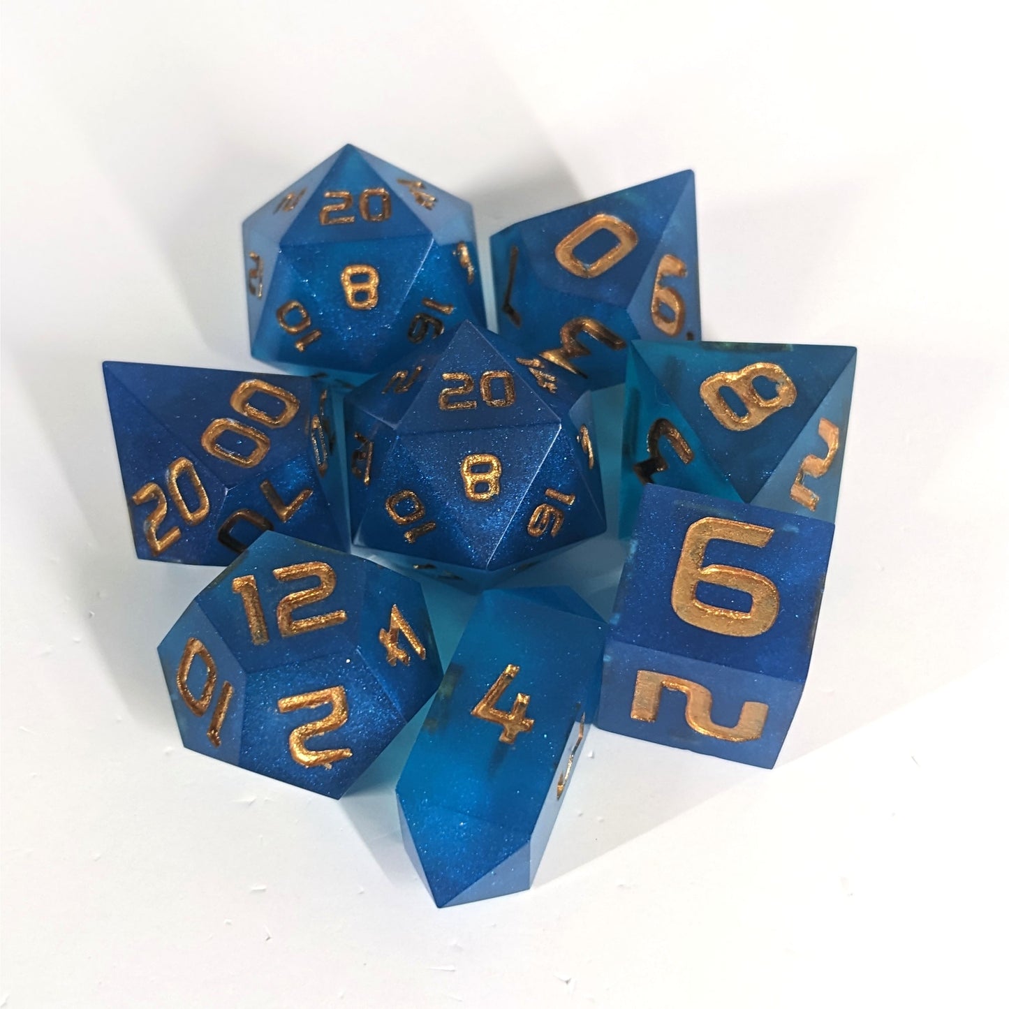 A complete set of 8 handmade artisan dice that are rich blue with gold numbers. The DnD dice set is arranged in a circle and features 2 D20s, one of which is in the center surrounded by the other 7 dice on a white background. The d4 is a unique, crystalline shape.