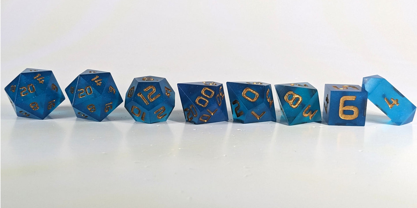 A straight on photo of a complete set of polyhedral dice that are rich blue with gold numbers arranged in a straight line. They are lined up with the 2 d20s on the left going down to the d4 on the right side. The highest value number of each die is facing forward, and the crystalline d4 is leaning against the d6.