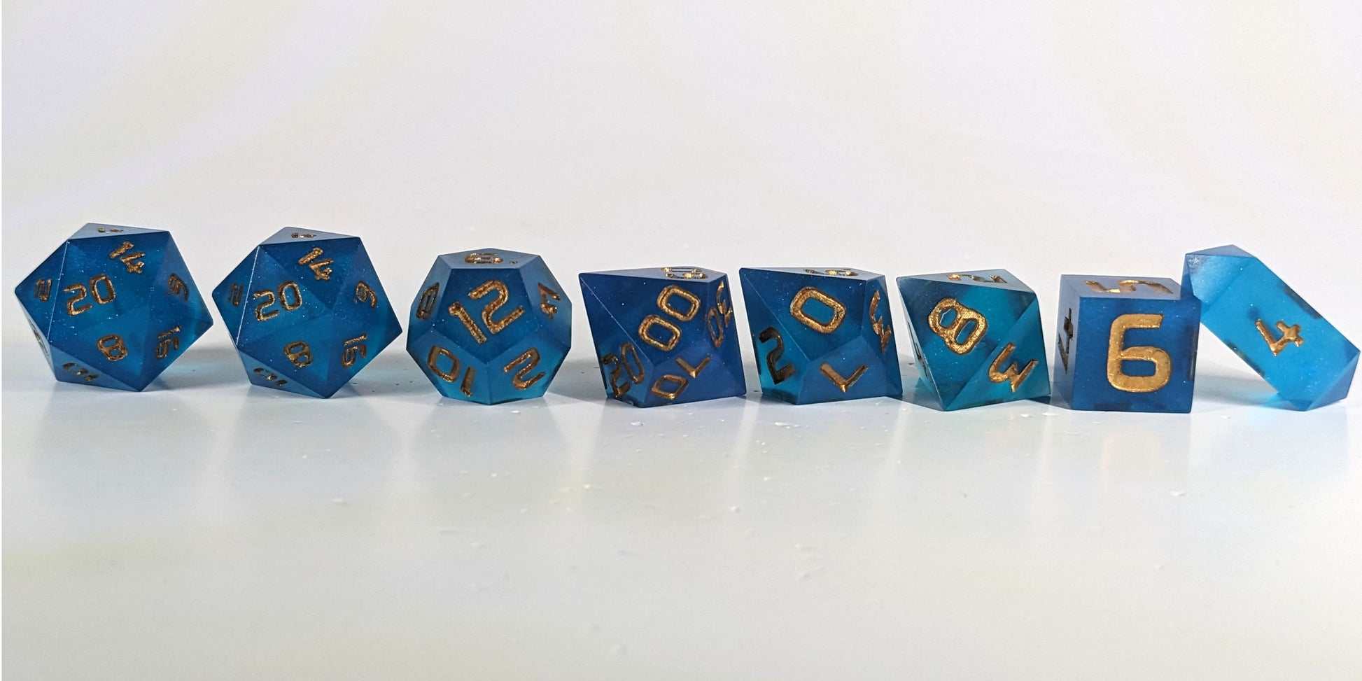 A straight on photo of a complete set of polyhedral dice that are rich blue with gold numbers arranged in a straight line. They are lined up with the 2 d20s on the left going down to the d4 on the right side. The highest value number of each die is facing forward, and the crystalline d4 is leaning against the d6.