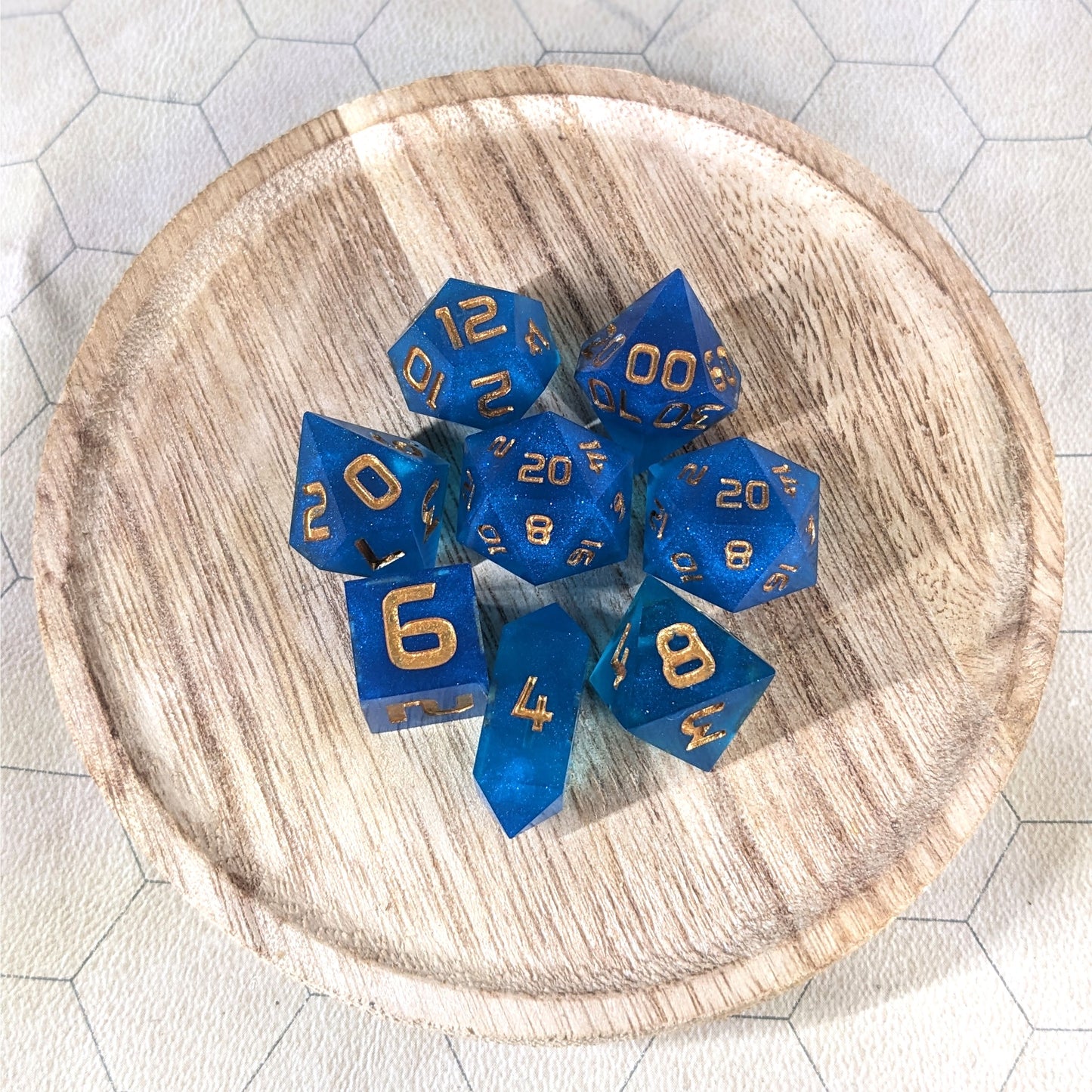 A complete set of 8 handmade artisan dice that are a rich blue with gold numbers. The DnD dice set is on a wooded circle and the dice are arranged in a circle and features 2 D20s, one of which is in the center surrounded by the other 7 dice. The d4 is unique with a crystalline shape.