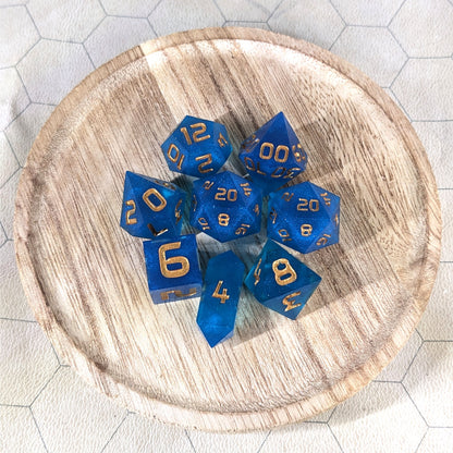 A complete set of 8 handmade artisan dice that are a rich blue with gold numbers. The DnD dice set is on a wooded circle and the dice are arranged in a circle and features 2 D20s, one of which is in the center surrounded by the other 7 dice. The d4 is unique with a crystalline shape.