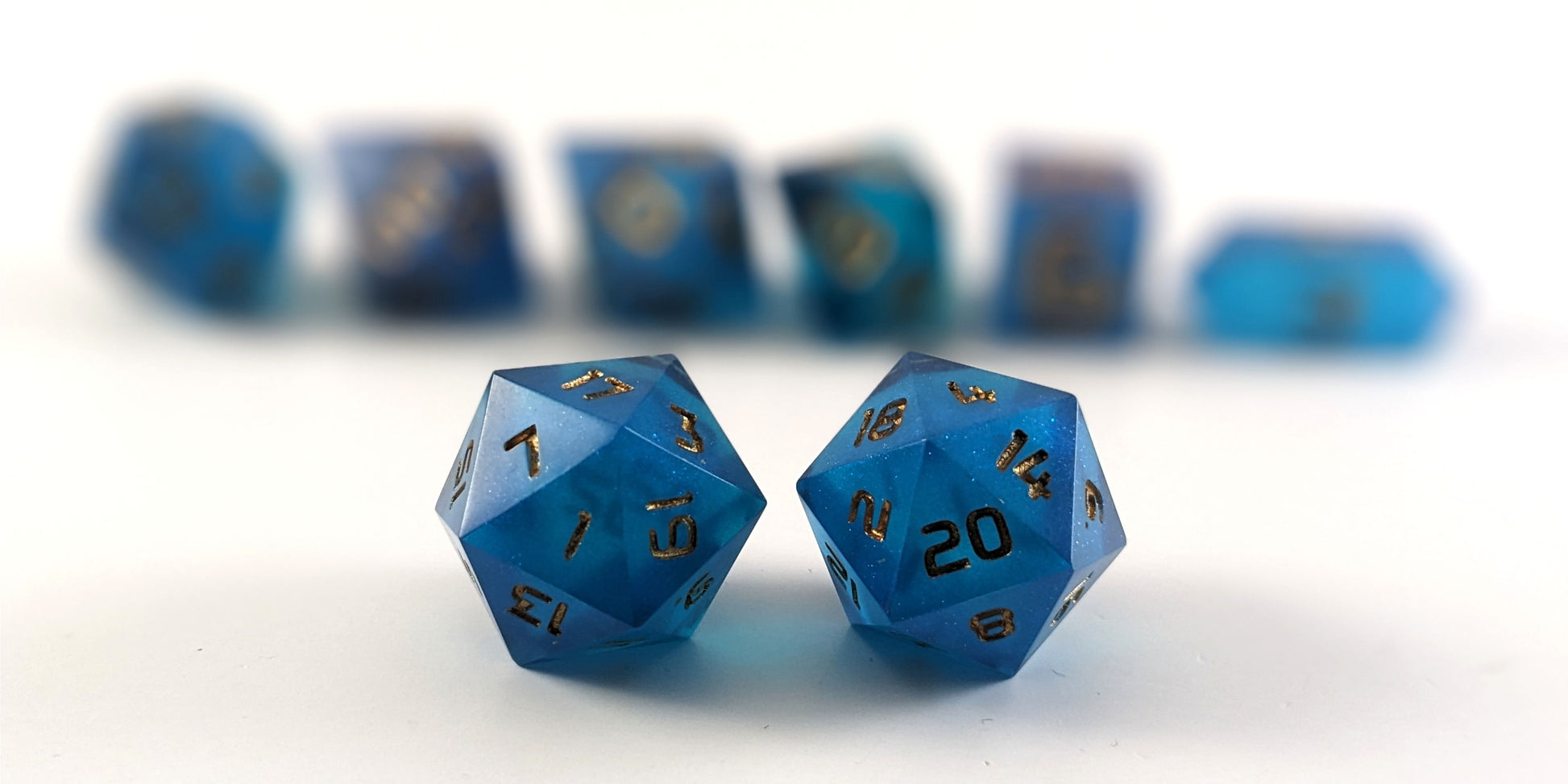A straight on photo of a standard set of DnD dice that are rich blue with gold numbers focusing on 2 d20s in the center. The d20 on the left has the 1 face centered and the d20 on the right has the 20 face centered. The other 6 dice are lined up in the background but are out of focus, while the d20s are in sharp focus.