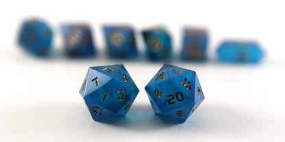A straight on photo of a standard set of DnD dice that are rich blue with gold numbers focusing on 2 d20s in the center. The d20 on the left has the 1 face centered and the d20 on the right has the 20 face centered. The other 6 dice are lined up in the background but are out of focus, while the d20s are in sharp focus.