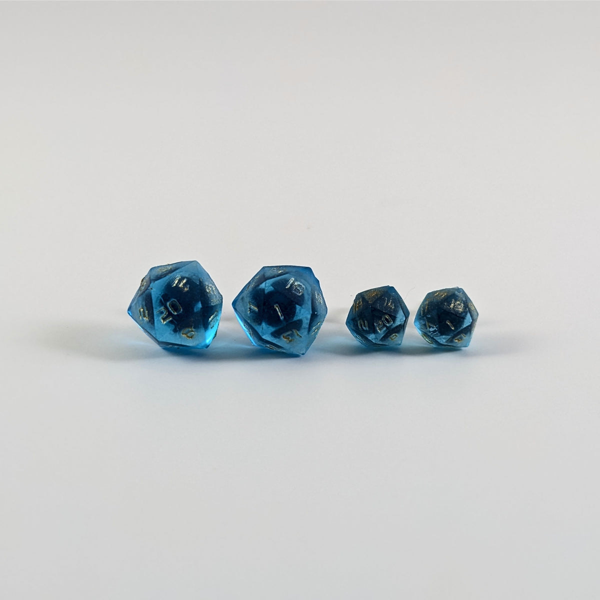 Two pairs of handmade d20 earrings with titanium hardware that are sapphire blue with gold numbers on a white background sit in a line with the larger pair on the left and the smaller pair on the right. The earrings on the left of each pair centers the 20 face and the earrings on the right of each pair centers the 1 face.