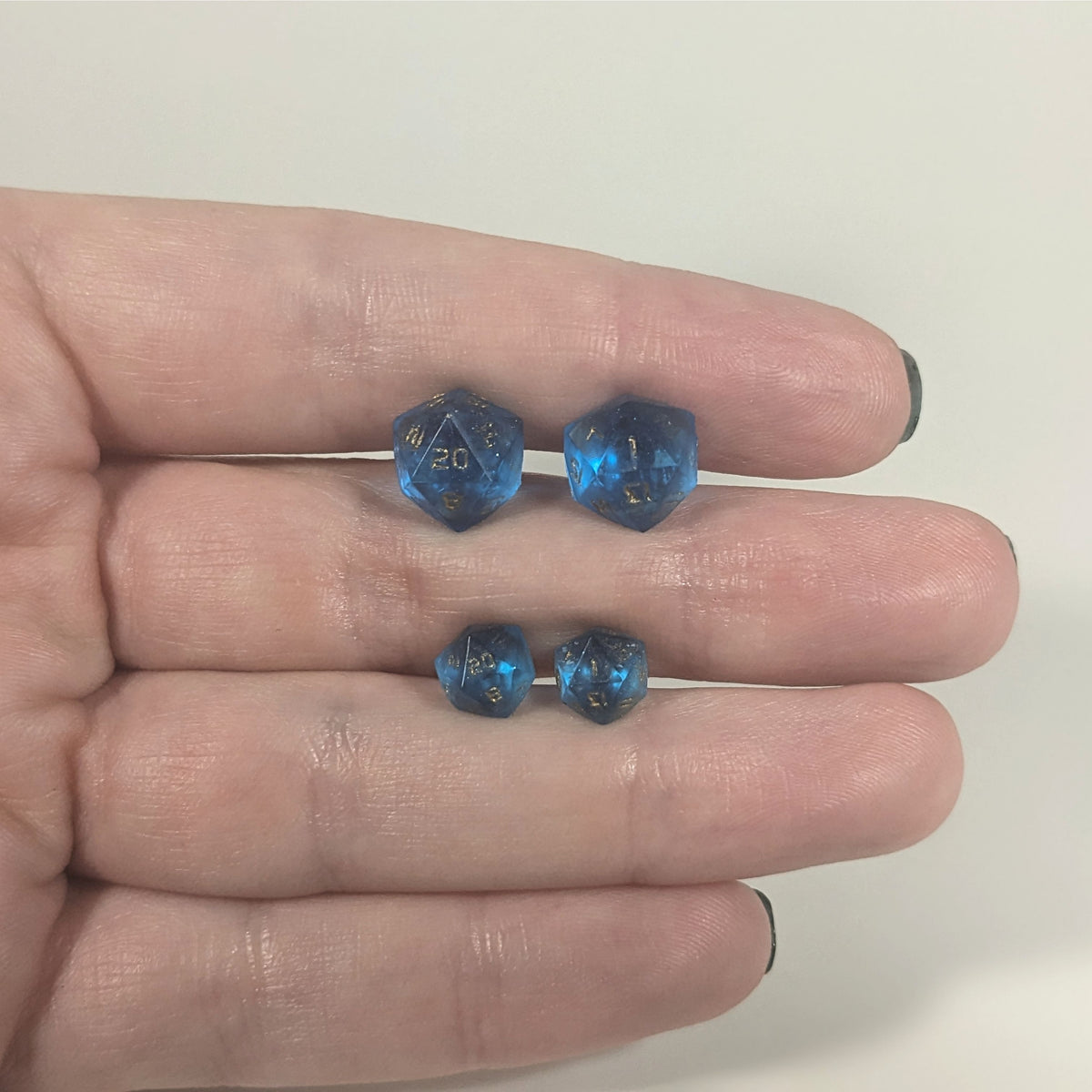 Two pairs of handmade d20 earrings with titanium hardware that are sapphire blue with gold numbers held between the fingers of a flat hand. The larger pair is between the pointer and middle finger, the smaller between the middle and ring finger. The earrings on the left of each pair centers the 20 face and the earrings on the right of each pair centers the 1 face.