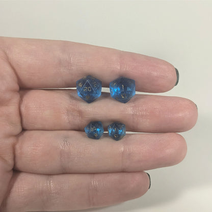 Two pairs of handmade d20 earrings with titanium hardware that are sapphire blue with gold numbers held between the fingers of a flat hand. The larger pair is between the pointer and middle finger, the smaller between the middle and ring finger. The earrings on the left of each pair centers the 20 face and the earrings on the right of each pair centers the 1 face.