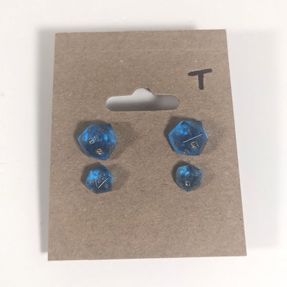 Two pairs of handmade d20 earrings with titanium hardware that are sapphire blue with gold numbers on a kraft brown earring card with a T in the top right corner. The earrings on the left of each pair centers the 20 face and the earrings on the right of each pair centers the 1 face.