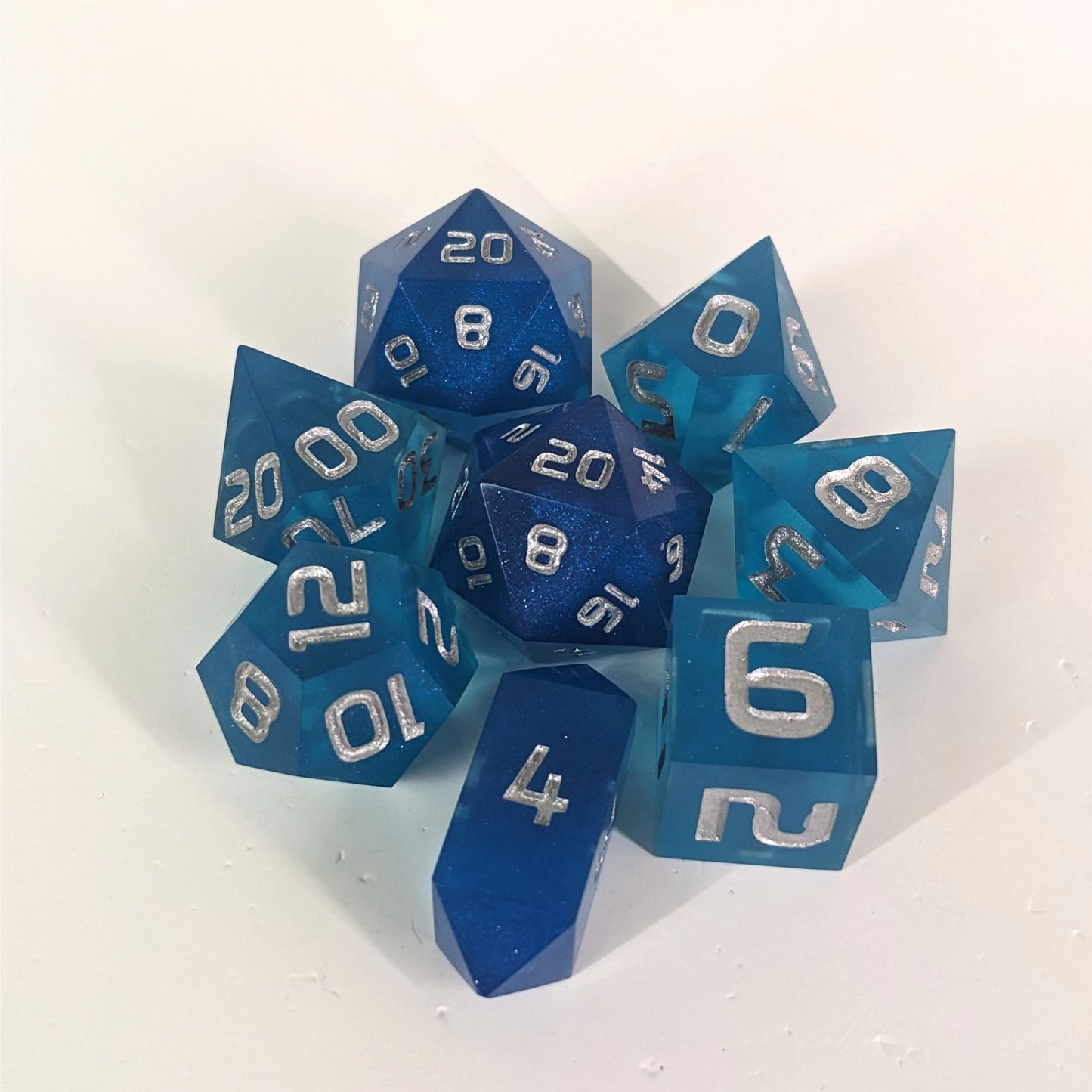 A complete set of 8 handmade artisan dice that are rich blue with silver numbers. The DnD dice set is arranged in a circle and features 2 D20s, one of which is in the center surrounded by the other 7 dice on a white background. The d4 is a unique, crystalline shape.