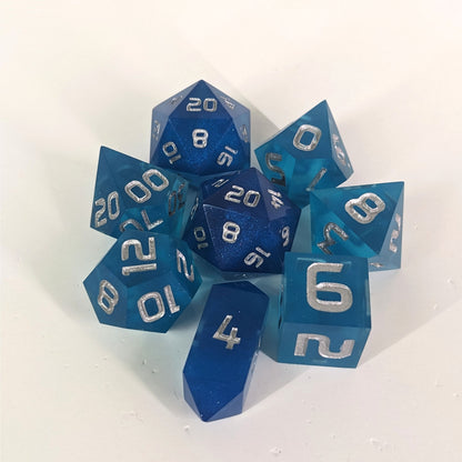 A complete set of 8 handmade artisan dice that are rich blue with silver numbers. The DnD dice set is arranged in a circle and features 2 D20s, one of which is in the center surrounded by the other 7 dice on a white background. The d4 is a unique, crystalline shape.