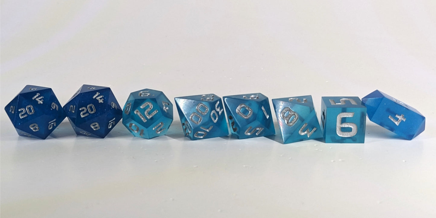 A straight on photo of a complete set of polyhedral dice that are rich blue with silver numbers arranged in a straight line. They are lined up with the 2 d20s on the left going down to the d4 on the right side. The highest value number of each die is facing forward, and the crystalline d4 is leaning against the d6.