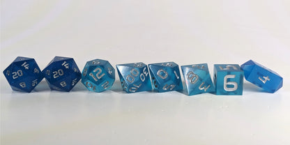 A straight on photo of a complete set of polyhedral dice that are rich blue with silver numbers arranged in a straight line. They are lined up with the 2 d20s on the left going down to the d4 on the right side. The highest value number of each die is facing forward, and the crystalline d4 is leaning against the d6.