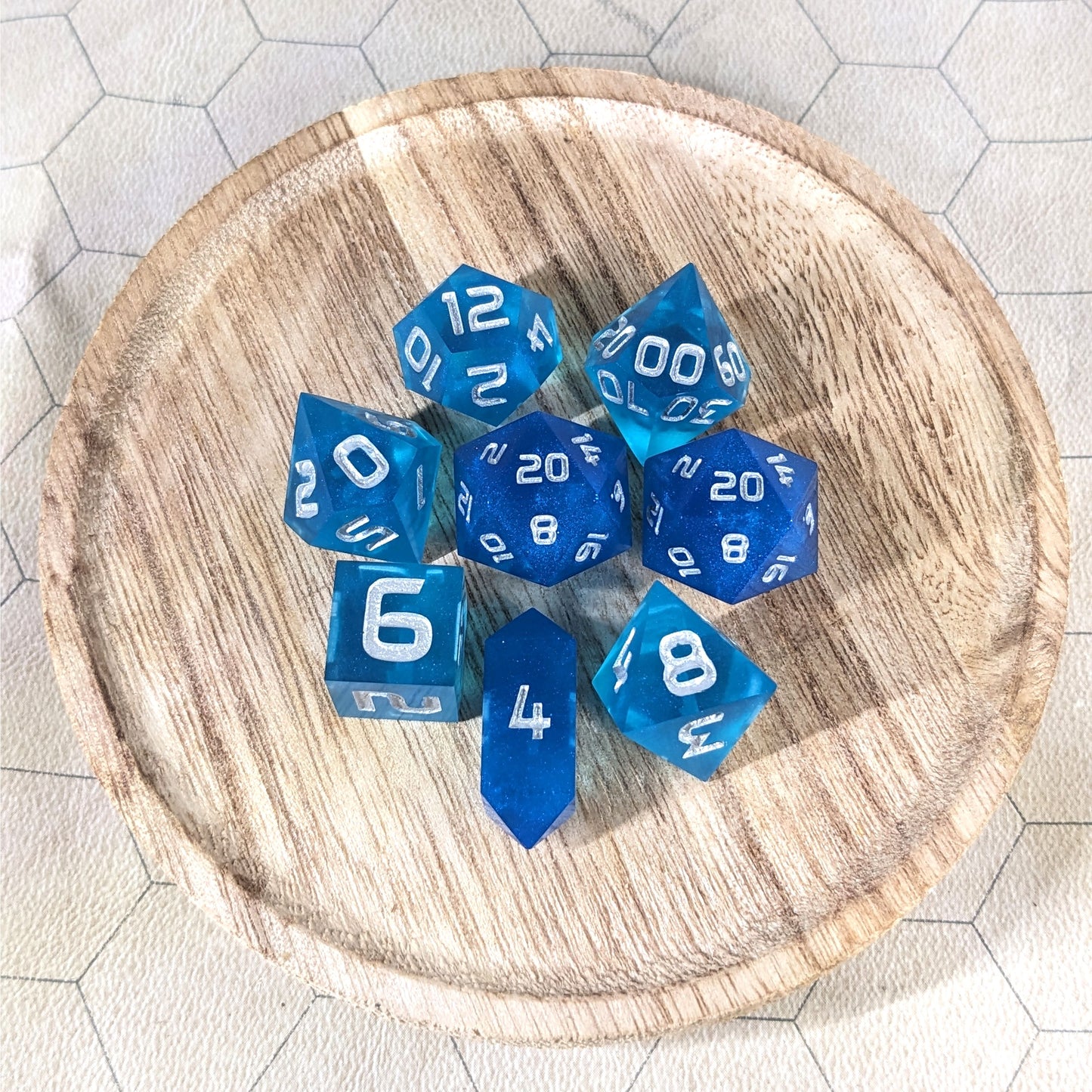 A complete set of 8 handmade artisan dice that are a rich blue with silver numbers. The DnD dice set is on a wooded circle and the dice are arranged in a circle and features 2 D20s, one of which is in the center surrounded by the other 7 dice. The d4 is unique with a crystalline shape.