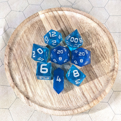 A complete set of 8 handmade artisan dice that are a rich blue with silver numbers. The DnD dice set is on a wooded circle and the dice are arranged in a circle and features 2 D20s, one of which is in the center surrounded by the other 7 dice. The d4 is unique with a crystalline shape.