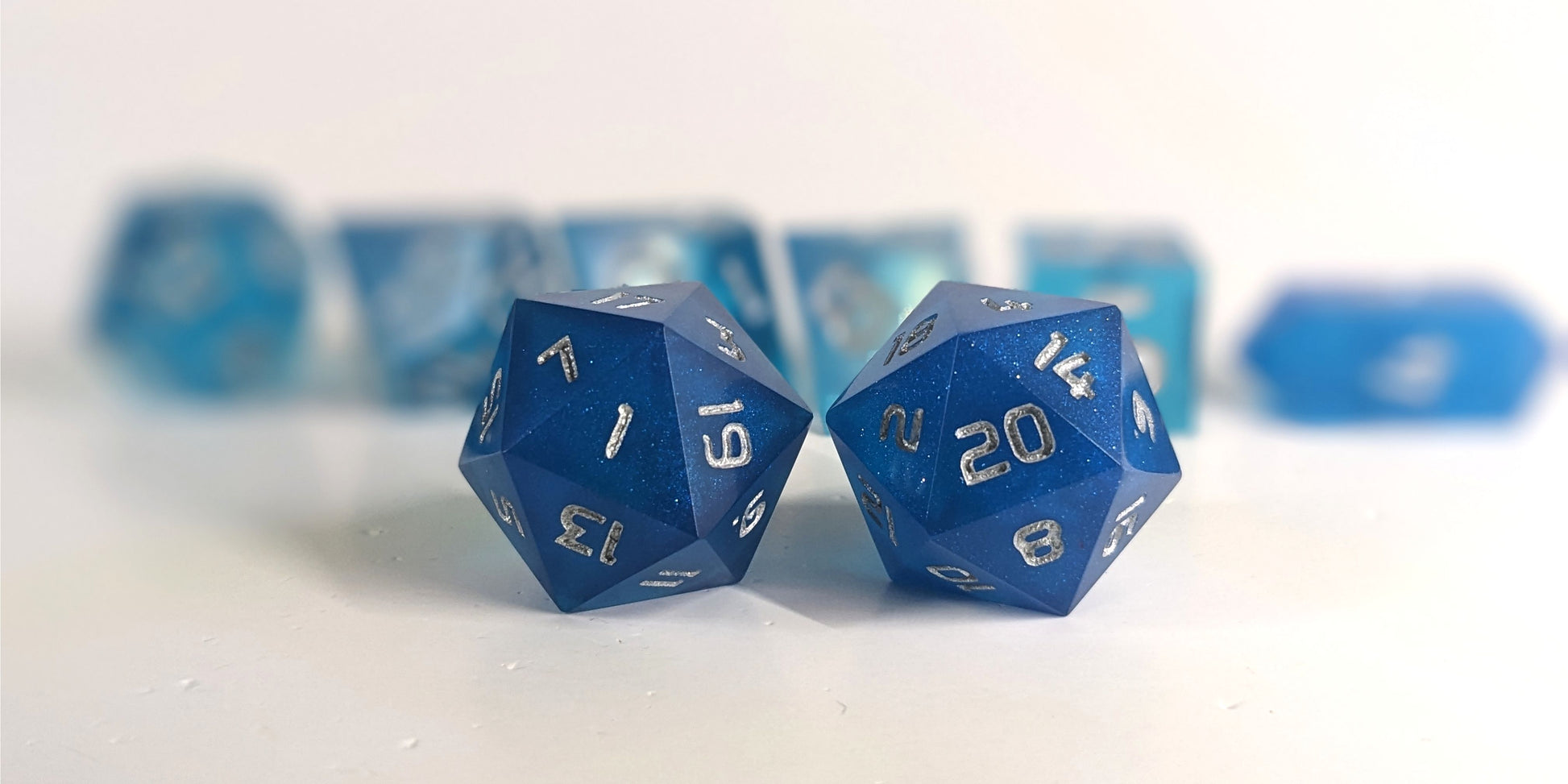 A straight on photo of a standard set of DnD dice that are rich blue with silver numbers focusing on 2 d20s in the center. The d20 on the left has the 1 face centered and the d20 on the right has the 20 face centered. The other 6 dice are lined up in the background but are out of focus, while the d20s are in sharp focus.
