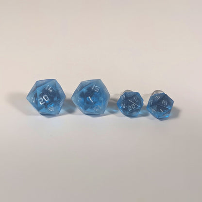 Two pairs of handmade d20 earrings with titanium hardware that are sapphire blue with silver numbers on a white background sit in a line with the larger pair on the left and the smaller pair on the right. The earrings on the left of each pair centers the 20 face and the earrings on the right of each pair centers the 1 face.