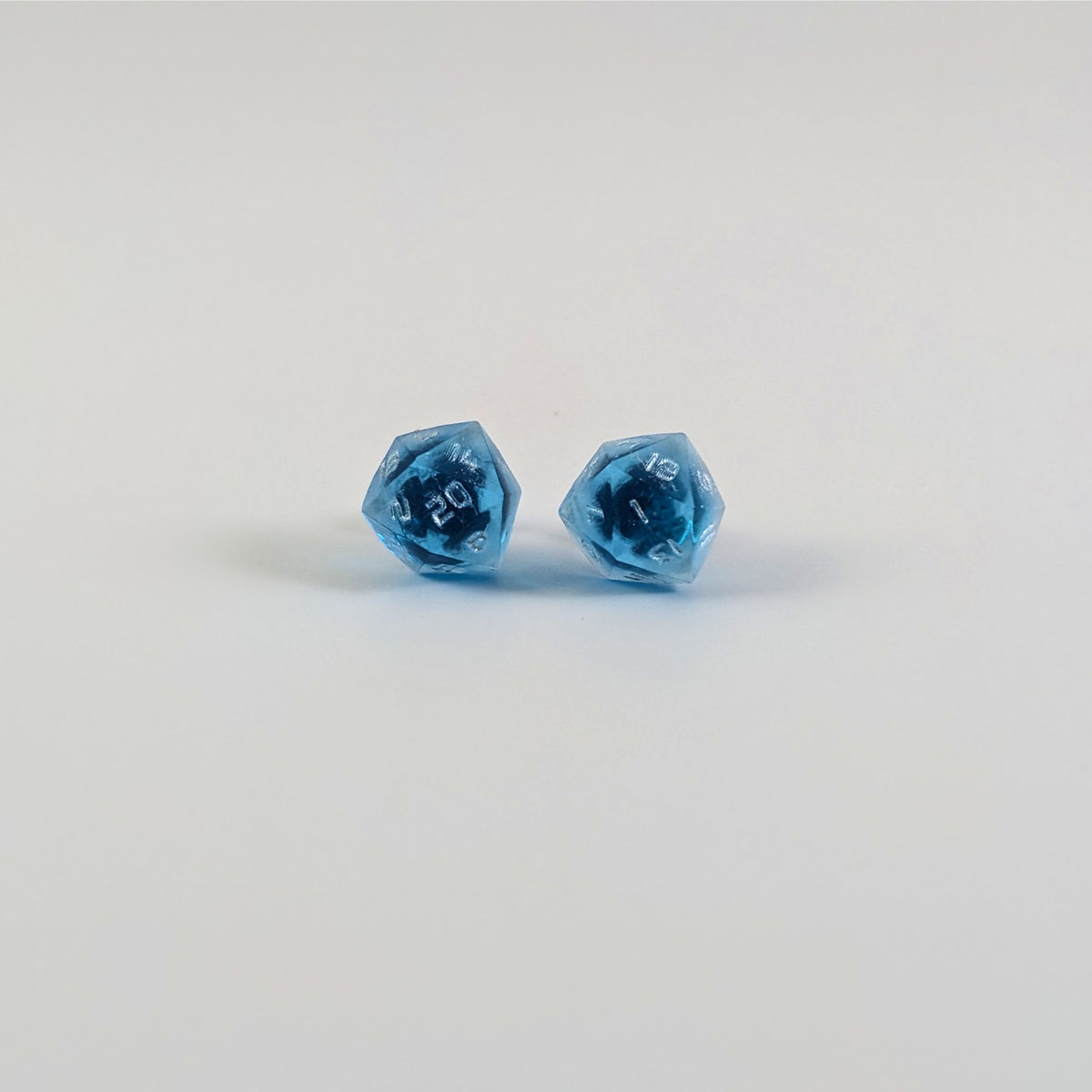 A single pair of the larger size handmade d20 earrings with titanium hardware that are sapphire blue with silver numbers on a white background. The earring on the left centers the 20 face and the earring on the right centers the 1 face.