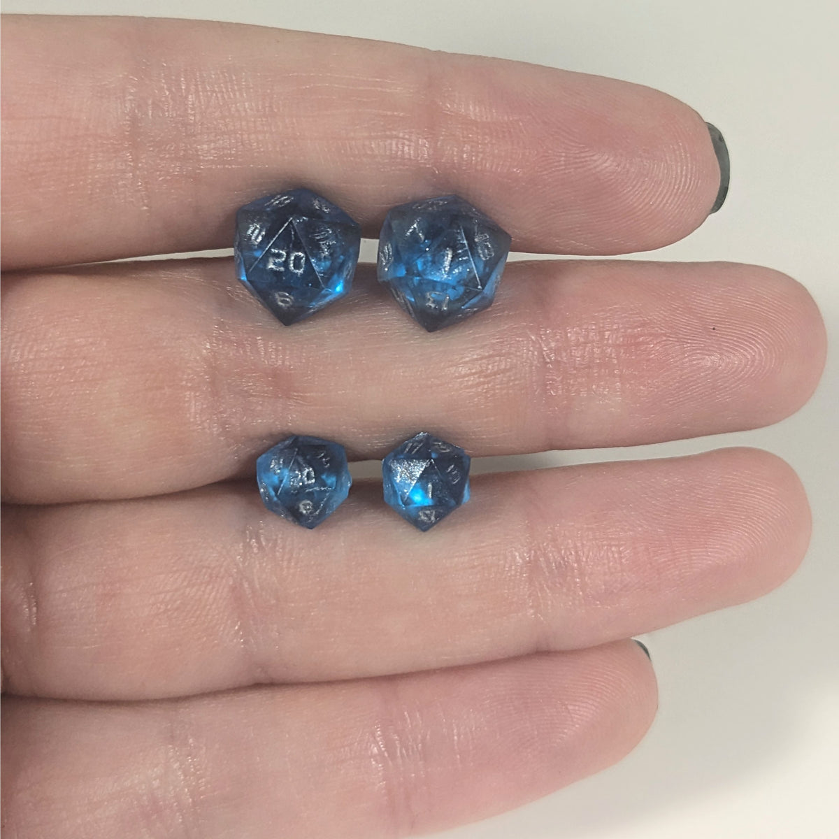 Two pairs of handmade d20 earrings with titanium hardware that are sapphire blue with silver numbers held between the fingers of a flat hand. The larger pair is between the pointer and middle finger, the smaller between the middle and ring finger. The earrings on the left of each pair centers the 20 face and the earrings on the right of each pair centers the 1 face.