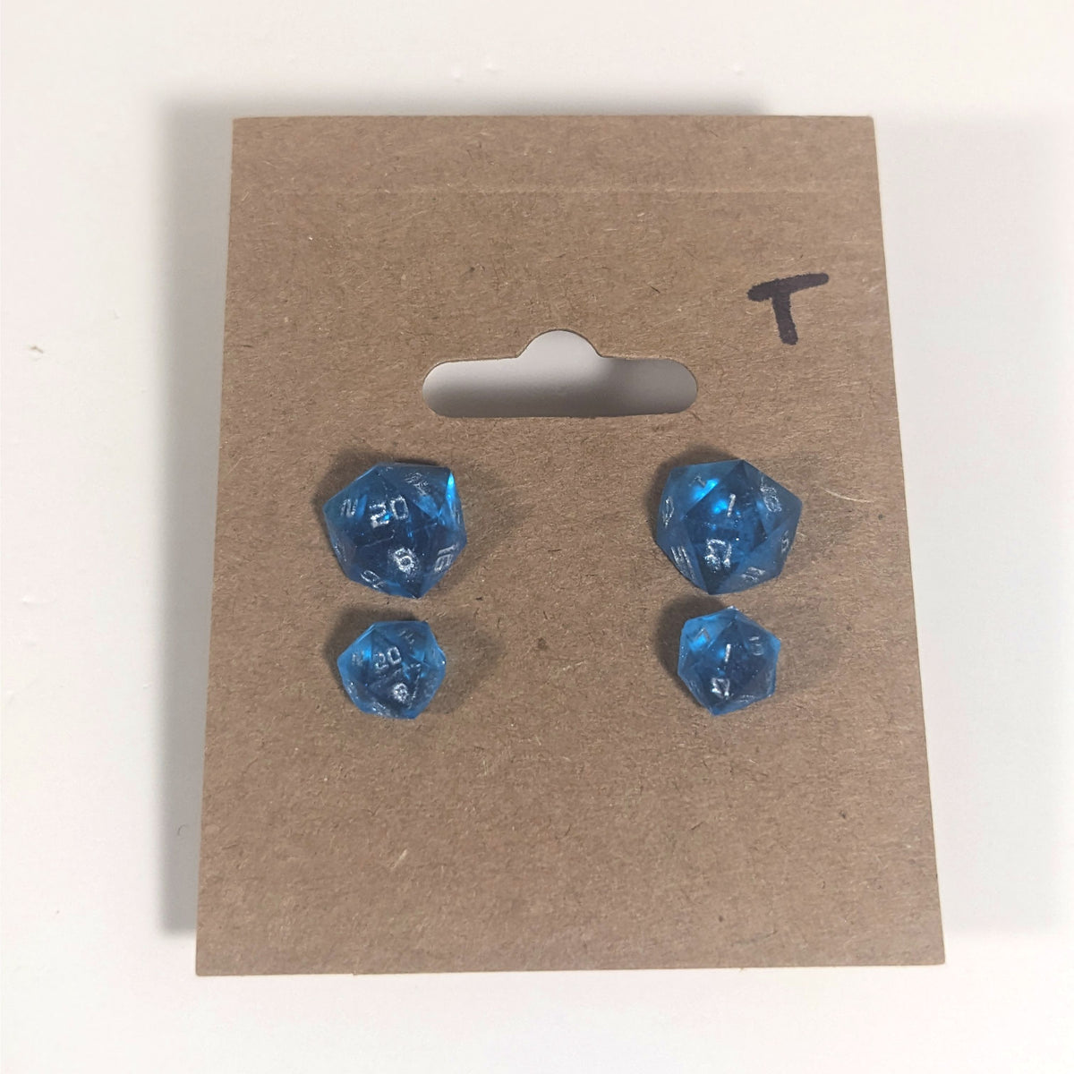 Two pairs of handmade d20 earrings with titanium hardware that are sapphire blue with silver numbers on a kraft brown earring card with a T in the top right corner. The earrings on the left of each pair centers the 20 face and the earrings on the right of each pair centers the 1 face.